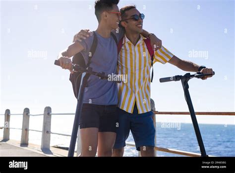 Happy Biracial Gay Male Couple Walking With Scooters Embracing On Promenade By The Sea Stock