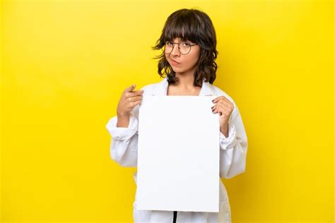 Jovem médica latina isolada em fundo amarelo segurando um cartaz vazio e apontando para a frente