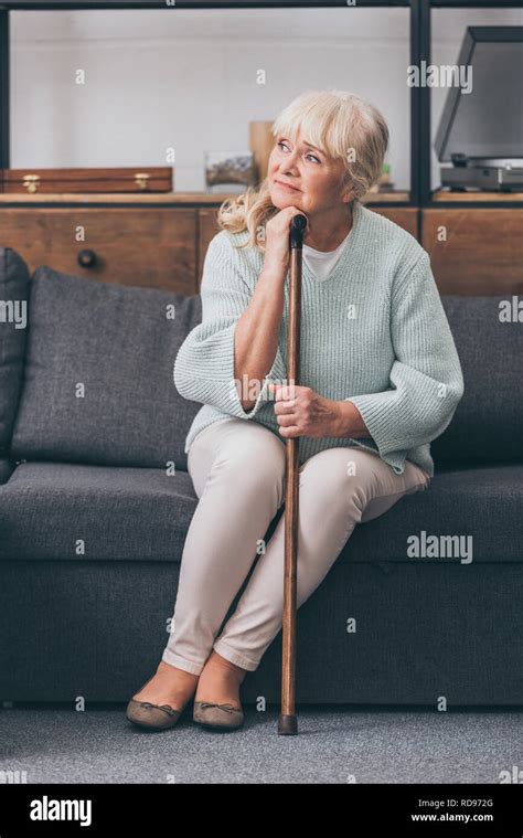 Upset Senior Woman With Walking Cane Sitting On Sofa At Home Stock