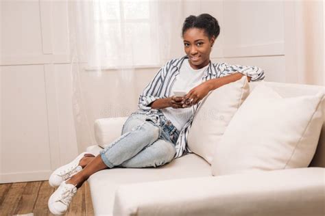 Pretty African American Woman Sitting On Couch And Using Smartphone