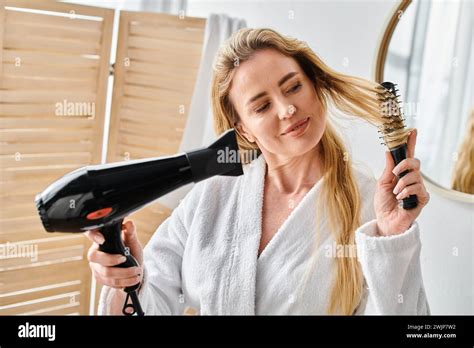 Good Looking Blonde Joyous Woman In Bathrobe Using Hair Dryer And Brush