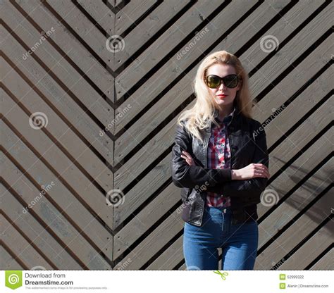 Beautiful Blonde Girl In Huge Sunglasses And A Black Jacket Posing Nex To Wooden Wall On A Sunny