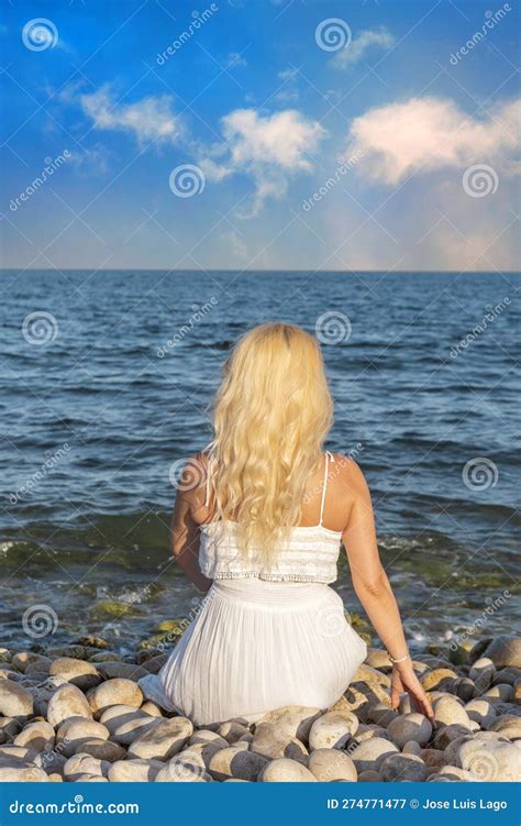 Femme Blonde En Robe Blanche Vue De Derrière Assise Sur Une Plage De Galets Face à La Mer Image