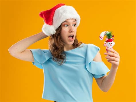 Free Photo Young Woman In Blue Top And Santa Hat Holding Christmas Candy Cane Looking At It