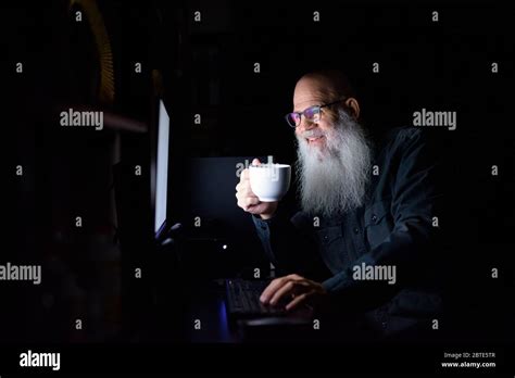 Happy Mature Bald Bearded Man Drinking Coffee While Working Overtime At
