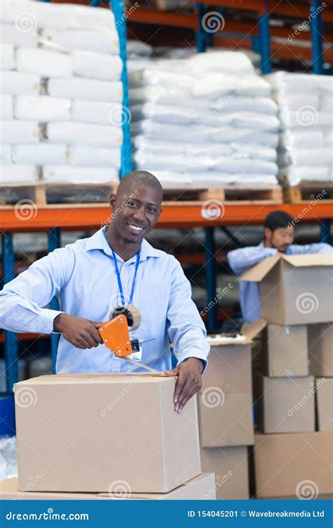 Male Staff Packing Cardboard Box With Tape Gun Dispenser In Warehouse