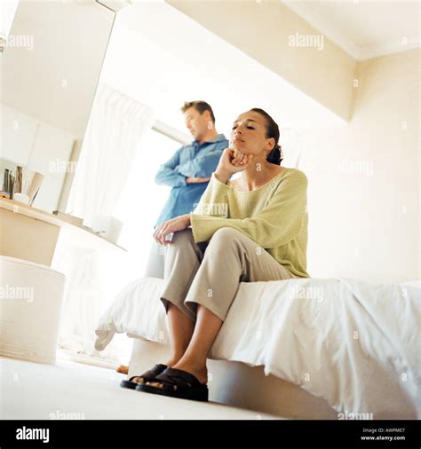 Man Standing With Arms Crossed Behind Woman Sitting On Edge Of Bed