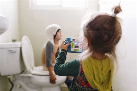 Young Girl Standing In Bathroom Holding Camera Taking Picture Of Boy Sitting On Toilet With