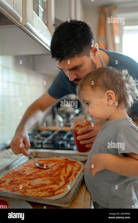 Boy By Father Spreading Tomato Sauce On Pizza Dough In Kitchen Stock