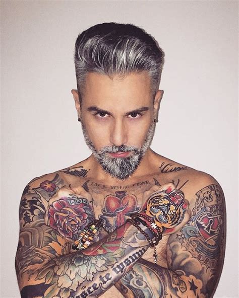 A Man With Grey Hair And Tattoos On His Chest Is Looking At The Camera While Holding His Arms