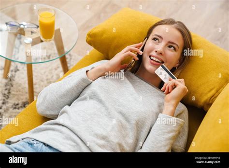 Schöne lächelnde junge blonde Frau in lässiger Kleidung auf einer Couch zu Hause ruhen zeigt