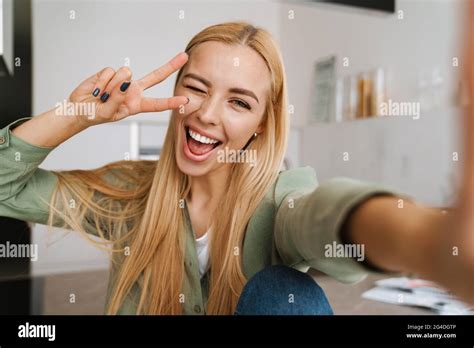 Happy Blonde Woman Showing Peace Sign While Taking Selfie Photo At Home