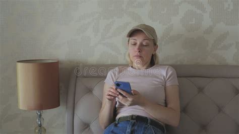 A Woman In A Baseball Cap Sitting On A Bed In A Hotel Room Uses A