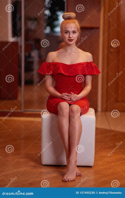 Une Jeune Belle Blonde En Robe Rouge Aux Pieds Nus Est Assise Sur Un Ottoman Blanc Dans Le Salon