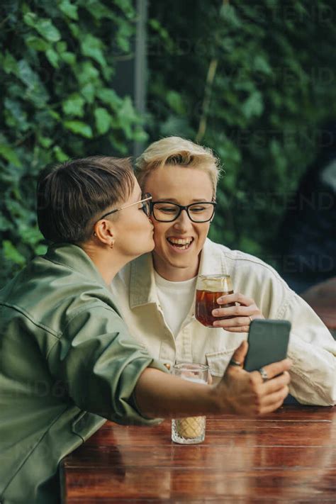 Smiling Lesbian Woman Kissing On Cheek Of Girlfriend And Taking Selfie Through Smart Phone Stock