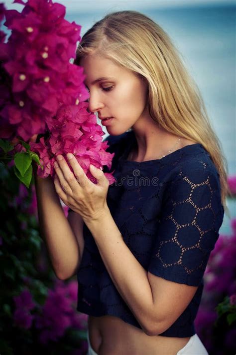 Blonde Caucasian Woman Wearing Dark Blue Lace Top And White Skirt Posing Between Pink Flowers