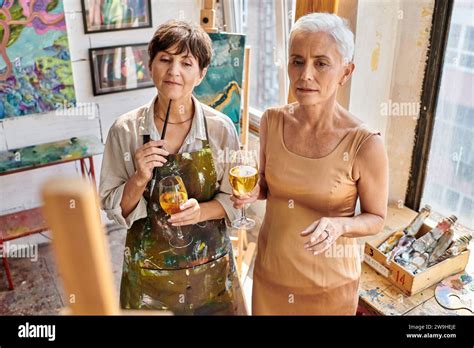 Thoughtful Woman Artist And Stylish Mature Model Standing With Wine Glasses In Craft Workshop