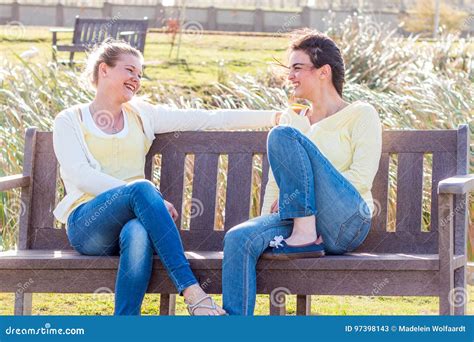 Two Happy Friends Sitting On Park Bench Talking And Interacting Stock