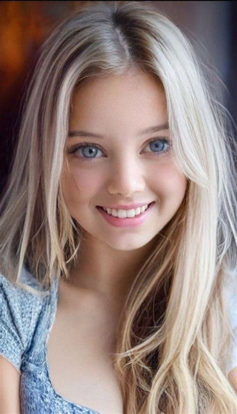 A Girl With Long Blonde Hair And Blue Eyes Smiling At The Camera While Wearing A Denim Shirt