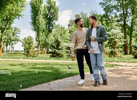 A Bearded Gay Couple Walks Hand In Hand Through A Green Park Enjoying