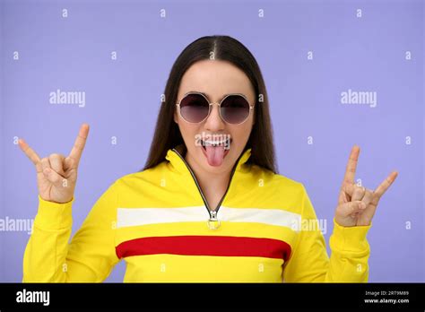 Happy Woman In Sunglasses Showing Her Tongue And Rock Gesture On Violet