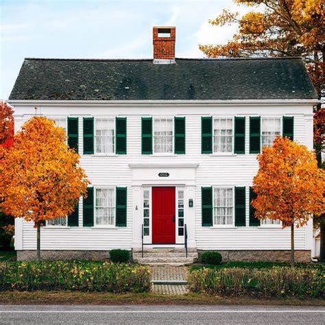 an old white house with red door and black shutters on the front, surrounded by autumn trees