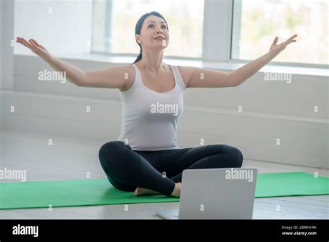 Brunette Female Sitting On Rug With Raised Hands Having Online Yoga
