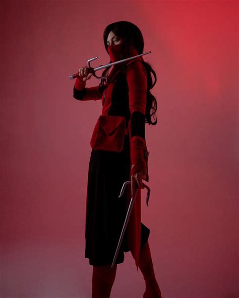 A Woman In A Red And Black Outfit Holding Two Swords While Standing