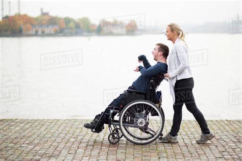 Caretaker Pushing Disabled Man On Wheelchair Along Lake Stock Photo