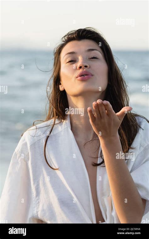 Brunette Woman In Shirt Blowing Air Kiss Near Blurred Sea Stock Photo