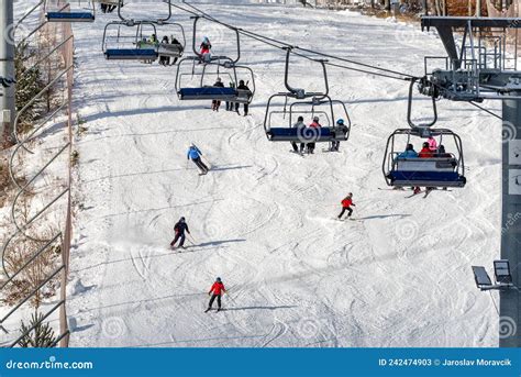 People Sitting On Chairlift And Skiers Skiing Downhill On Slope At