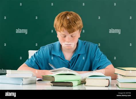 Boy In Classroom With Books On Desk Against Green Chalkboard Stock