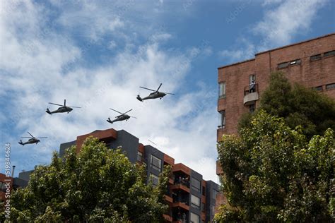 Four Black Hawk Helicopters Flying Over Two Brick Residential Buildings With Blue And Cloudy Sky