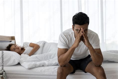 Upset Depressed Young Man Sitting On The Edge Of Bed Against His Wife