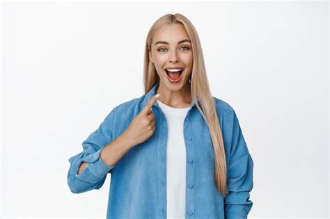 Fille blonde surprise et heureuse pointant le doigt sur elle même souriante et semblant excitée