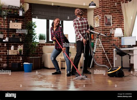 Woman Mopping The Floor And Dancing While Man Sings With Passion Holding Vacuum Cleaner African