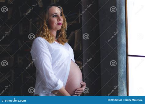 Happy Pregnant Woman Stands Near The Window And Hugs Her Belly With Her