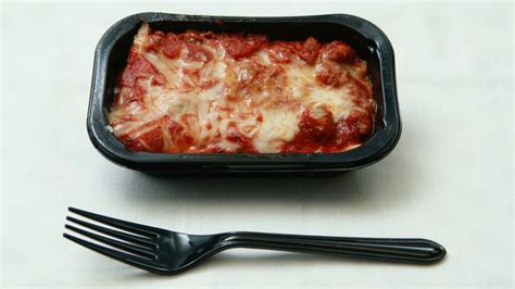 A Plastic Container Filled With Lasagna Next To A Fork On Top Of A Table