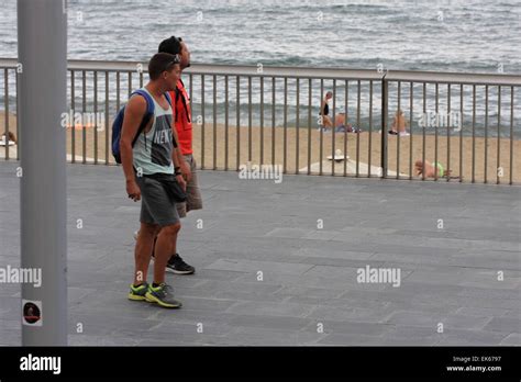 Two Male Tourists Walk Along The Mediterranean Sea Shore In Barcelona