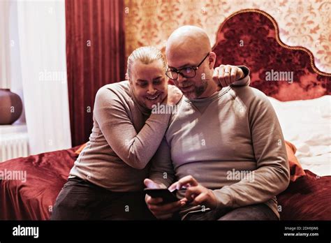 Gay Couple Looking At Mobile Phone While Sitting On Bed In Hotel Room