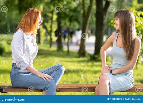 Two Young Girls Friends Sitting On A Bench In Summer Park Chatting