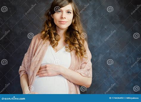 Portrait Of Beautiful Pregnant Caucasian Girl Touching Her Belly And Standing Over Black Wall