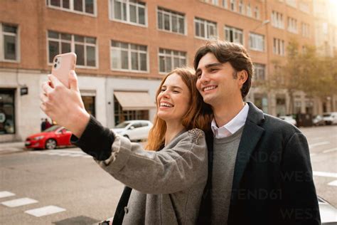 Happy Young Man Embracing Girlfriend Taking Selfie On Smartphone While