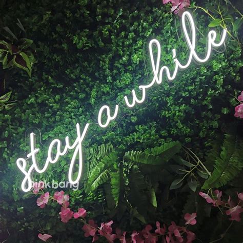 A Neon Sign That Says Stay Awhile In Front Of Some Plants And Flowers