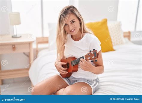 Young Blonde Woman Playing Ukulele Sitting On Bed At Bedroom Stock