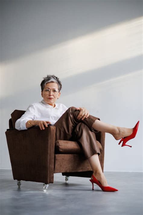 Fashionable Mature Woman In Red Heels Classic White Shirt And Eyeglasses Sitting On Armchair On