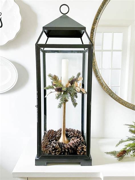 A Christmas Centerpiece With Pine Cones And Greenery In A Glass Lantern