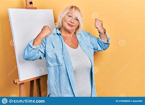 Middle Age Blonde Woman Standing By Painter Easel Stand Showing Arms