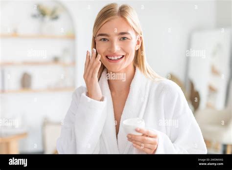 Blonde Woman Smiling To Camera While Applies Moisturizer In Bathroom Stock Photo Alamy