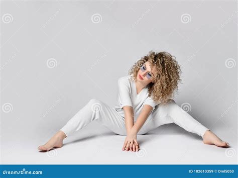 Blonde Krullende Meid In Blauw Zonnebril Wit Pak En Barefoot Zit Op De Vloer Met Haar Benen